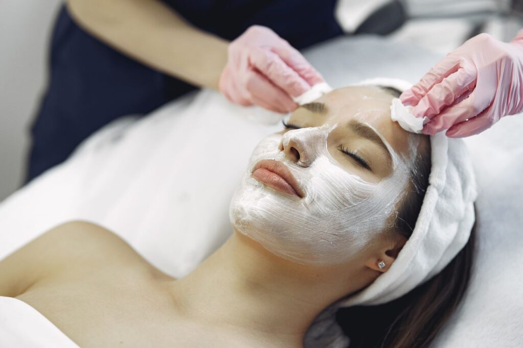 From above anonymous medical specialist in uniform and gloves wiping cream mask off face of relaxed woman during skincare procedure in modern studio